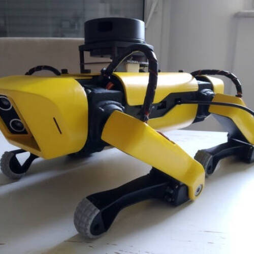 A yellow, four-legged robotic dog stands on a white table. The robot features a camera and sensors on its front and top, with black and yellow limbs and thick, textured feet. Cables and components are visible along its body.